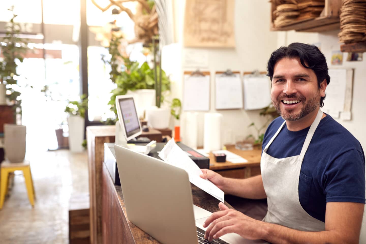 Portrait Of Male Sales Assistant Working On Laptop Behind Sales Desk Of Florists Store